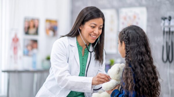 An OLE Health provider listens to a young patient's teddy bear with a stethoscope